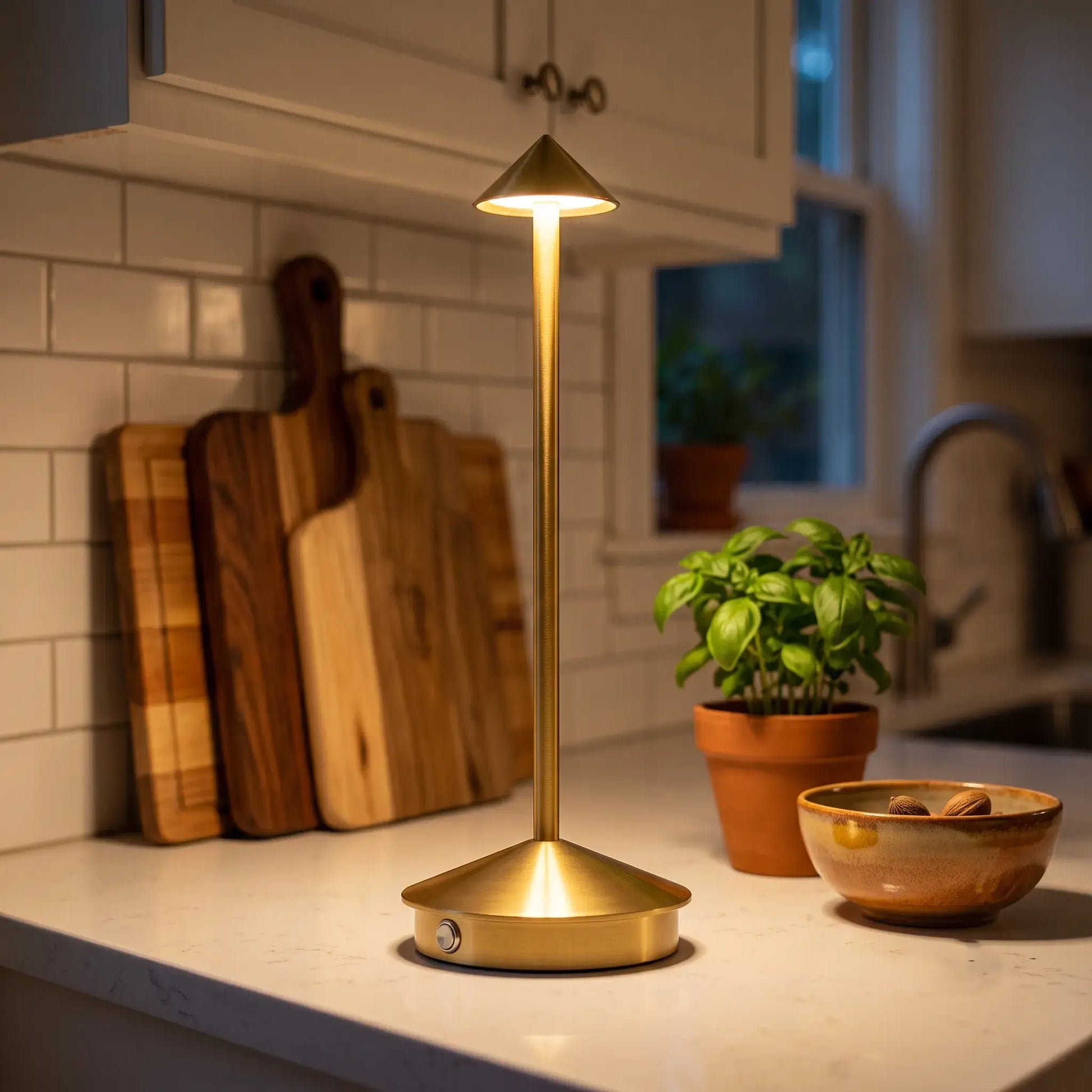 Aesthix Lume Vela gold slim rechargeable lamp on a kitchen countertop next to a potted basil plant and a ceramic bowl.