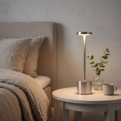 Aesthix Lume Halo silver cordless dimmable table lamp glowing beside pillows on a bedside table