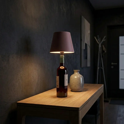 Aesthix Lume Vino, A wine bottle lamp shaped like a dark wine bottle with a  pink fabric shade, illuminated and placed on a light wood table next to a ceramic vase in a dimly lit interior hallway.